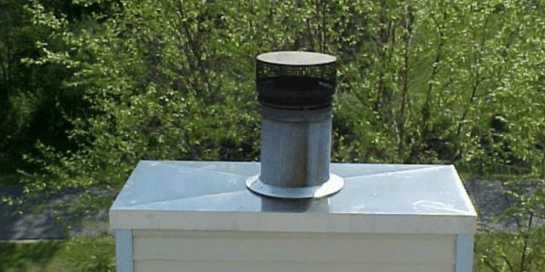 A metal chimney pipe extends from the top of a white roof, surrounded by greenery and trees in the background, indicating recent roofing repairs. The chimney has a cylindrical cap on top.